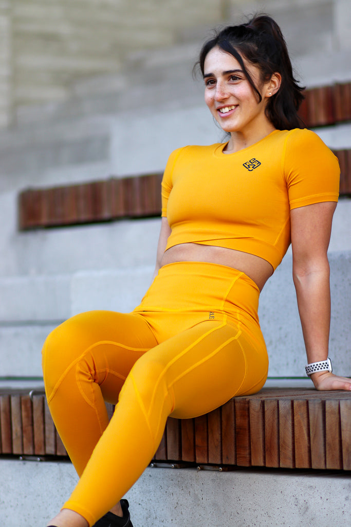 Happy athlete sitting on a bench outdoors. She is wearing a yellow crop top and high waisted leggings from Agu Athletics.