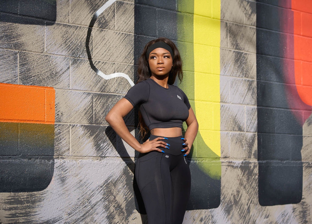 Woman standing in front of mural modelling the black yogas set from Agu Athletics. The set consists of a fitted crop top and high-waisted, squat proof leggings.