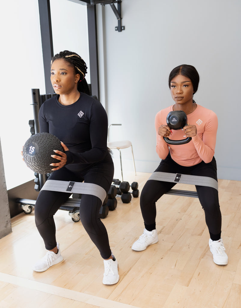 Two girls exercising in the gym with the Agu Athletics Hip Bands. Also, they are wearing the Fearless long sleeve gym shirt. One in black, the other in rose.
